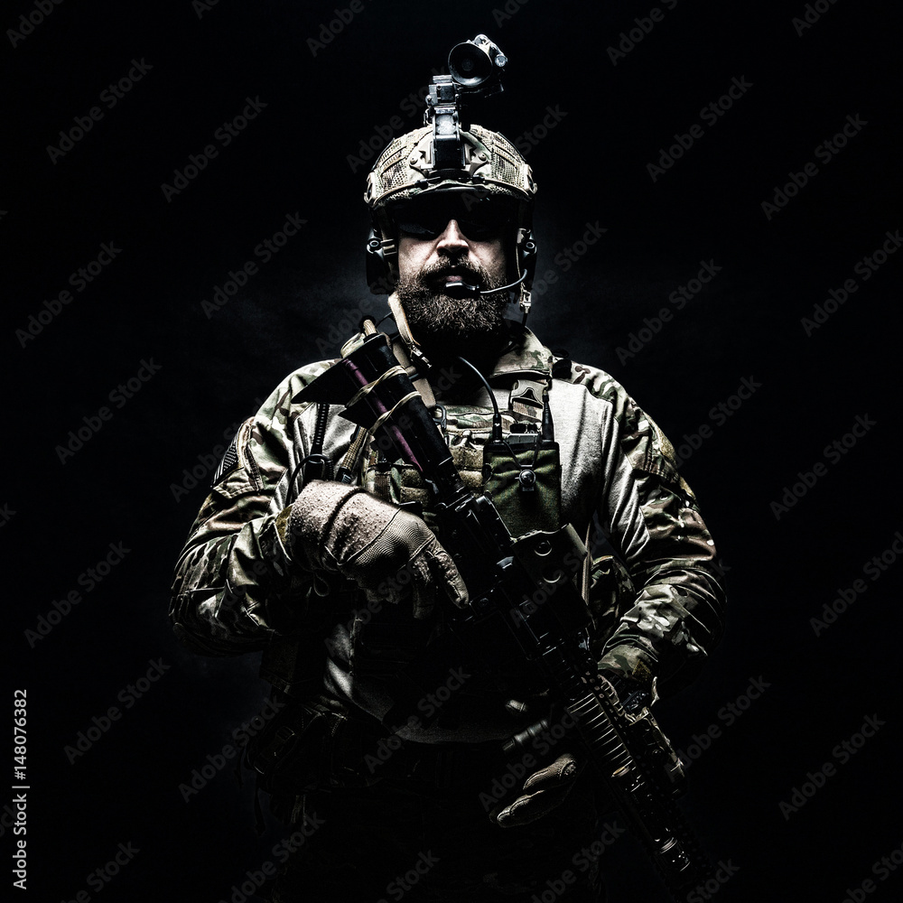Bearded soldier in Combat Uniforms with weapon, plate carrier and combat helmet are on. Studio shot, dark background. Ruthless and fearless, he brings death to enemies, defending his country