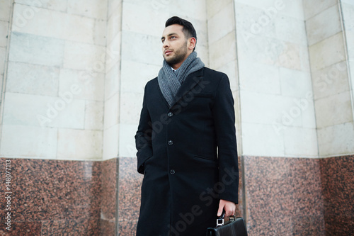 Portrait of handsome successful businessman standing against marble wall of building and looking away confidently