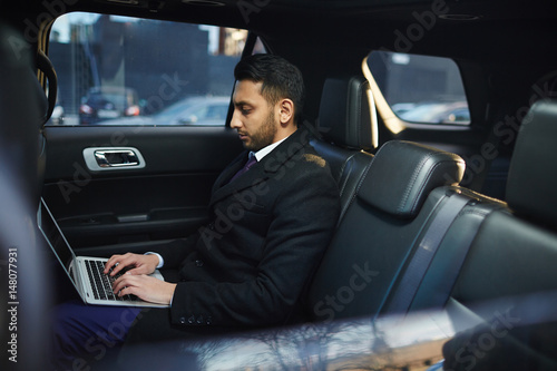 Side view portrait of handsome Middle-Eastern businessman using laptop on backseat inside expensive car © pressmaster
