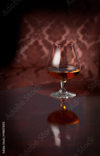 Glass of brandy, cognac or whiskey standing on a reflective table in front of patterned chair