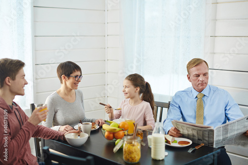 Close-knit family of four enjoying delicious breakfast at home: they sitting at kitchen table, eating cornflakes and chatting with each other