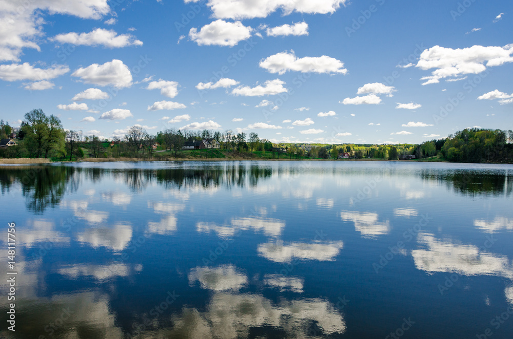 Cloudy blue sky reflects into the lake with trees on the horizon.