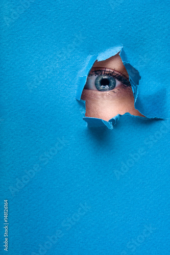 A female eye looks through a hole in a paper blue background.