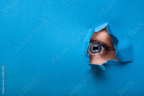 A female eye looks through a hole in a paper blue background.