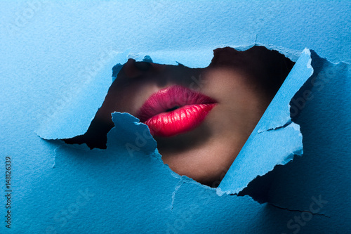 A view of the female lips through a hole in a paper blue background.