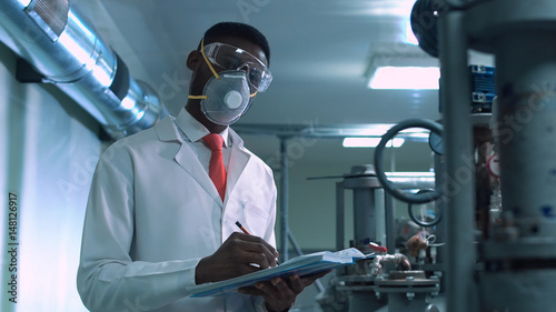 Side view of black scientist in white lab coat, goggles and mask standing next to productional equipment with journal and writing photo