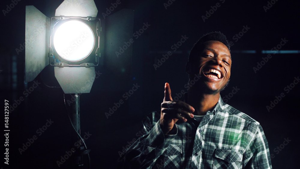 Naklejka premium Young African-American man in darkness coming close to camera, then studio light stand is being turned on making man happy and smiling. Looking at camera