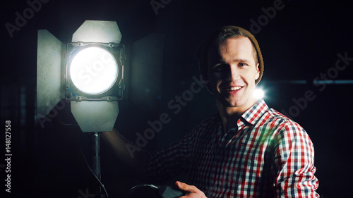 Stylish man in hat and standing at studio light stand with switcher in hand, turning it on and checking and smiling, looking at camera photo