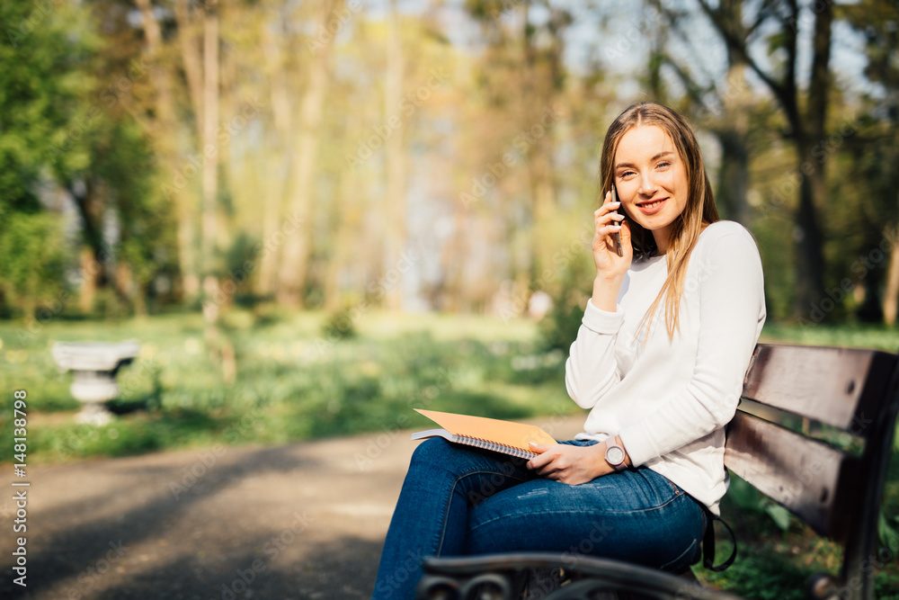 Portrait of attractive young professional woman using a smartphone while sitting on a wooden bench in a park, smiling. Portrait of attractive young professional woman using a smartphone while sitting on a wooden bench in a park, smiling.