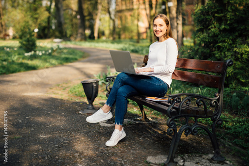 Happy young student with a tablet and a disposable coffee cup sitting on the bench and reading in a summer park.