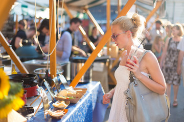 Beautiful blonde caucasian lady wearing white summer dress buying freshly prepared meal at a local street food festival. Urban international kitchen event in Ljubljana, Slovenia, in summertime.