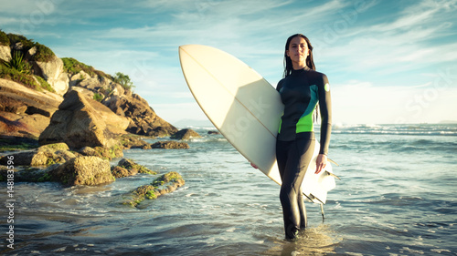 Young female surfer in ocean with surfboard at rocky beach