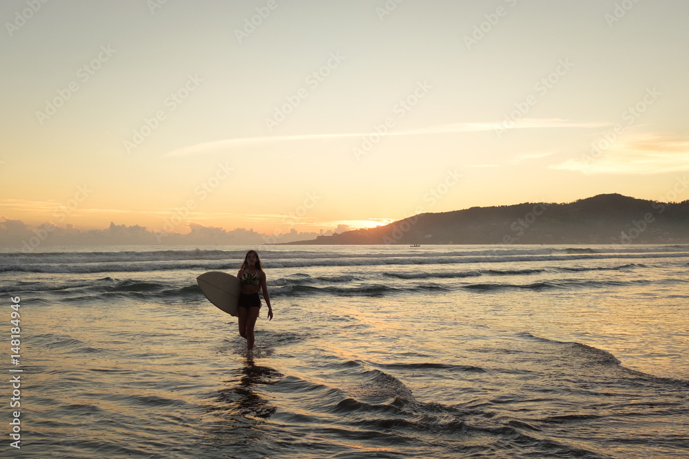Female surfer leaving the ocean with surfboard at sunset or sunrise