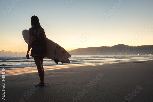 Female surfer with surfboard looking into the ocean during sunset sunrise