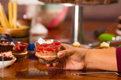 Hand grabbing onto delicious mousse covered in strawberry topping, presented inside small glass, cake pieces and muffins background, pastry concept