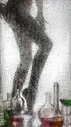 Legs of a beautiful naked girl behind a wet glass and with a still-life from bottles and wine glasses.