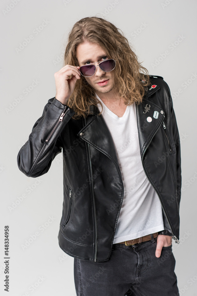young handsome rocker posing in black leather jacket and sunglasses isolated on grey young handsome rocker posing in black leather jacket and sunglasses isolated on grey