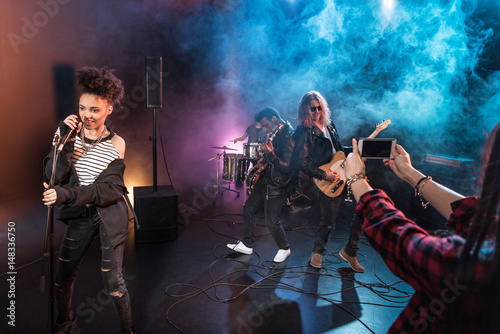 Young woman photographing rock and roll band performing hard rock music on stage