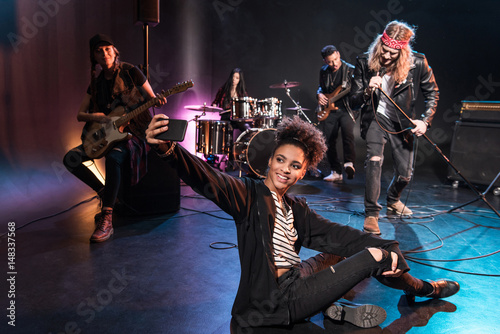 Young woman taking selfie with rock and roll band performing concert on stage