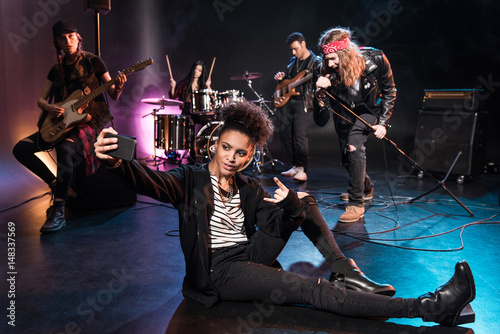 Young woman taking selfie with rock and roll band performing concert on stage