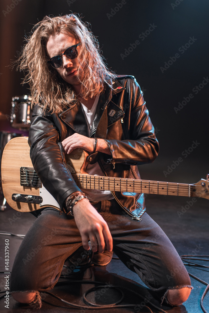 Fototapeta premium Handsome rock star in eyeglasses posing with electric guitar on stage