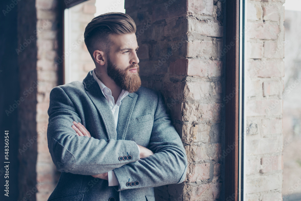 Close up portrait of handsome bearded young man in suit leaning on brick wall and looking in the window