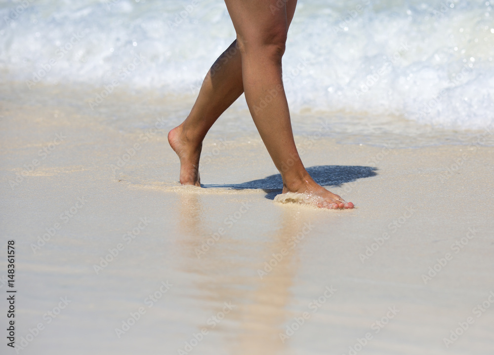 Caucasian tanned woman is walking on the beach. Water and sand on the feet. White sand beach with waves.
