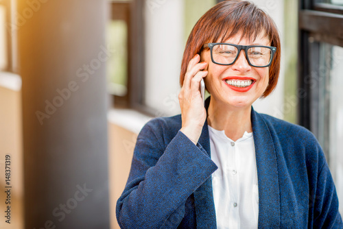 Older and elegant businesswoman talking phone standing indoors near the window