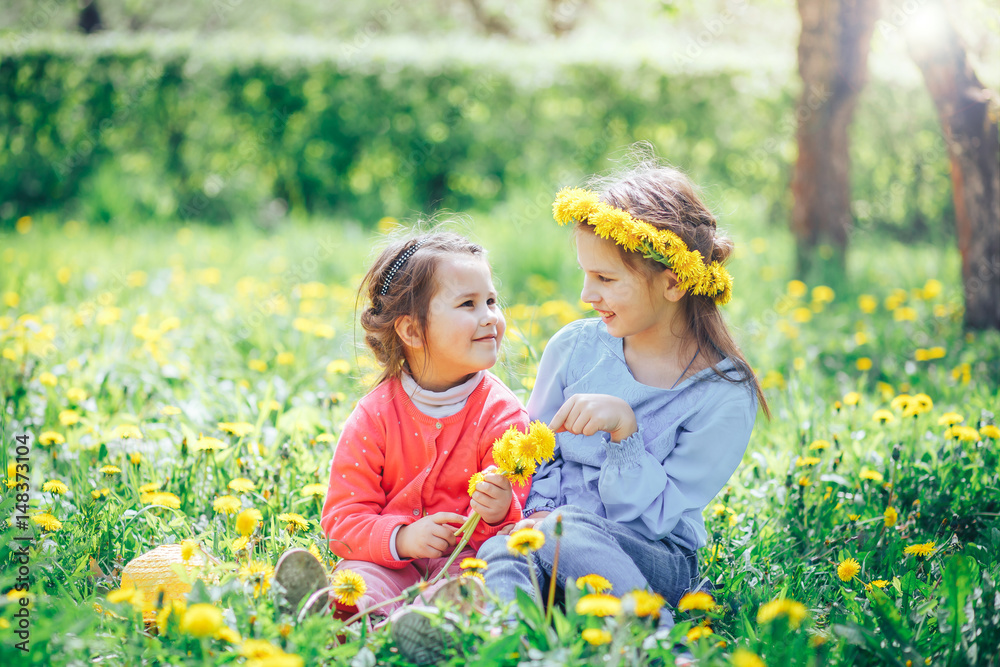 two beautiful little girls Caucasian and Asian appearance sitting on a green meadow and weave a wreath of dandelions two beautiful little girls Caucasian and Asian appearance sitting on a green meadow and weave a wreath of dandelions