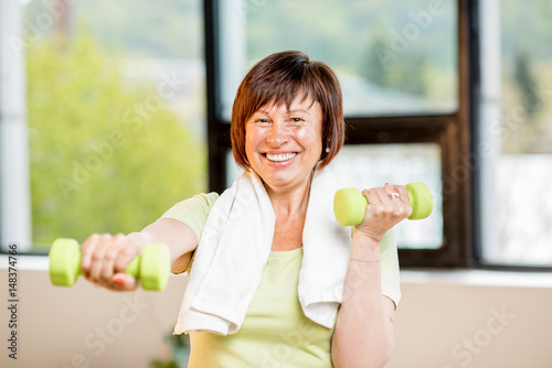 Happy older woman in sports wear training with dumbbells indoors on the window background