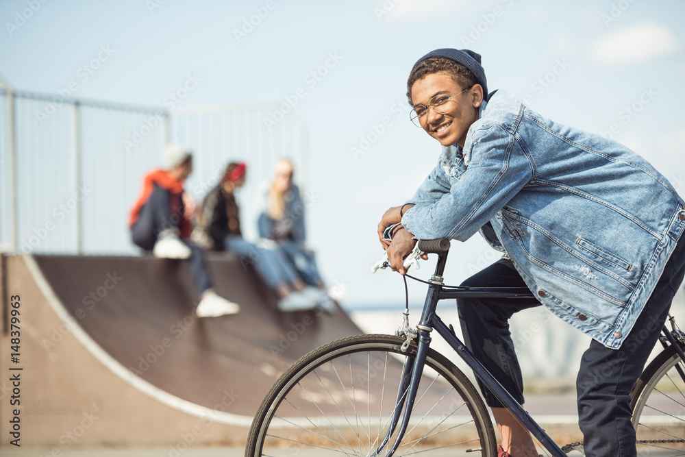 smiling hipster teenage boy riding bicycle at skateboard park
