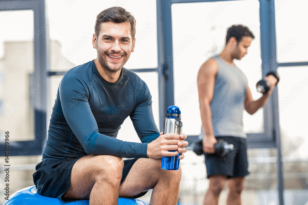 Handsome young man with bottle of water resting at gym, other man workout on blurred background