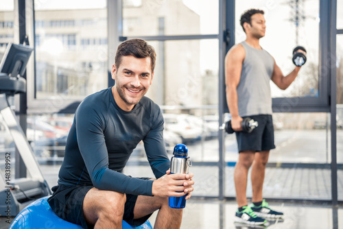 Handsome young man with bottle of water resting at gym  other man workout on blurred background