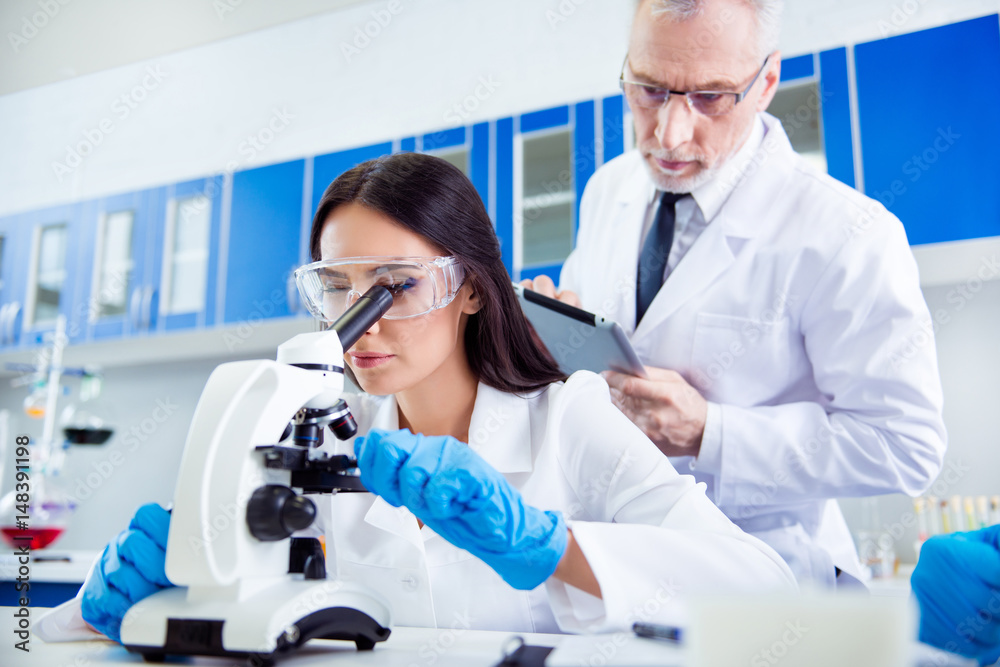 Laboratory, biotechnology, team work. Researcher is checking the esample on microscope and older professor is controlling her work and making notes. They are wearing labcoats Laboratory, biotechnology, team work. Researcher is checking the esample on microscope and older professor is controlling her work and making notes. They are wearing labcoats