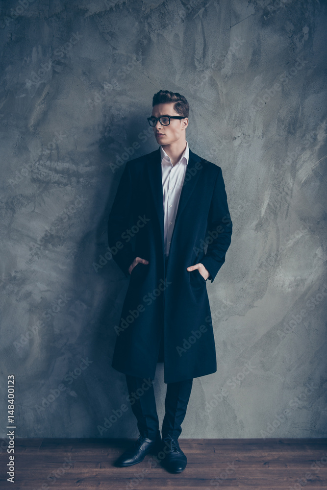Young attractive businessman in glasses put his arms into pockets of black coat. He is standing near the grey wall Young attractive businessman in glasses put his arms into pockets of black coat. He is standing near the grey wall