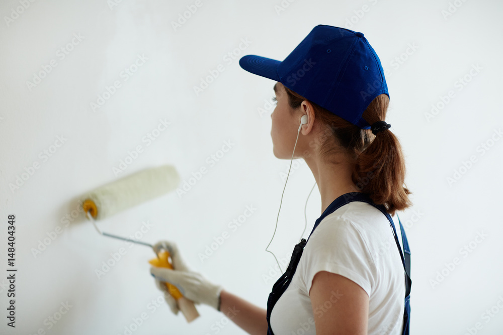 Rear view portrait of young woman on construction site: female worker painting walls white using paint roller while remodeling office and listening to music Rear view portrait of young woman on construction site: female worker painting walls white using paint roller while remodeling office and listening to music