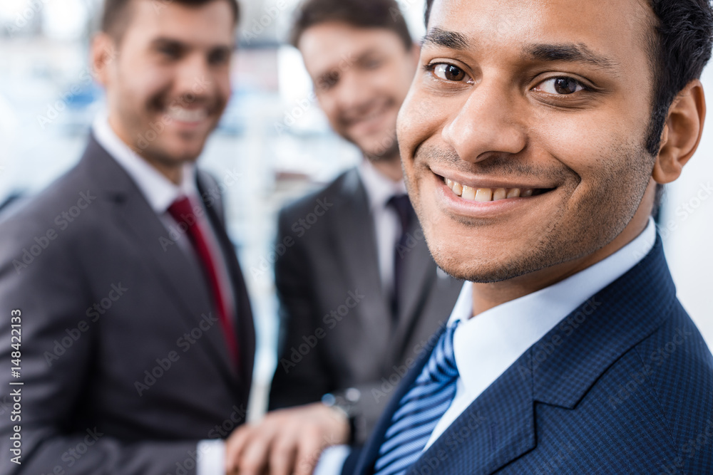 Close-up portrait of smiling african american businessman looking at camera with colleagues standing behind, business concept Close-up portrait of smiling african american businessman looking at camera with colleagues standing behind, business concept