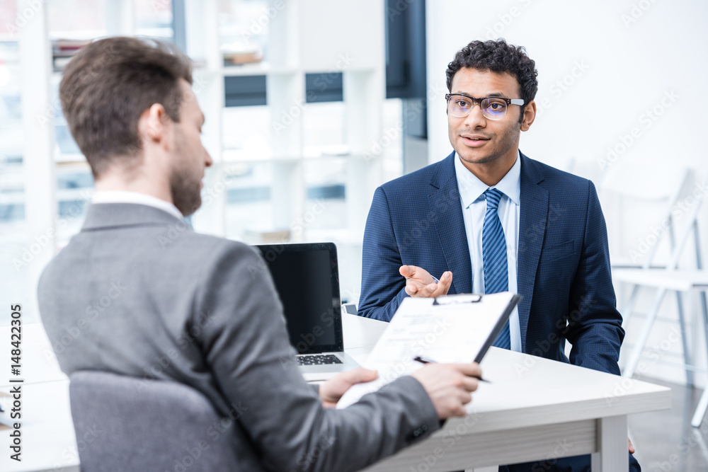 Smiling man in suit and eyeglasses gesturing and looking at businessman with clipboard, business concept Smiling man in suit and eyeglasses gesturing and looking at businessman with clipboard, business concept