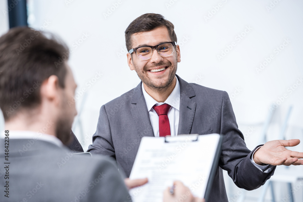 Smiling man in suit and eyeglasses gesturing and looking at businessman with clipboard, business concept Smiling man in suit and eyeglasses gesturing and looking at businessman with clipboard, business concept