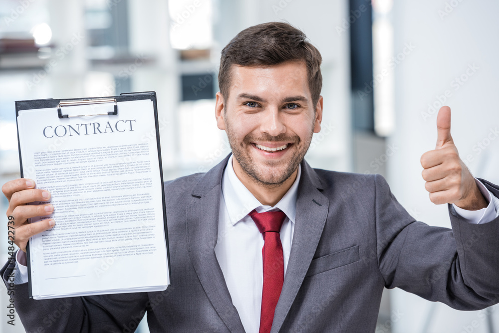 Handsome smiling businessman holding contract and showing thumb up in office, business concept Handsome smiling businessman holding contract and showing thumb up in office, business concept