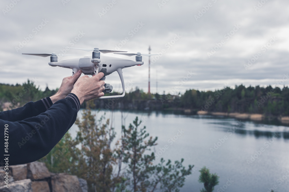 The man in dark pants and a jacket holding quadrocopter in his hands before takeoff on the background of a forest lake. Unmanned aerial copter. Aeromodelling, hobby, leisure concept