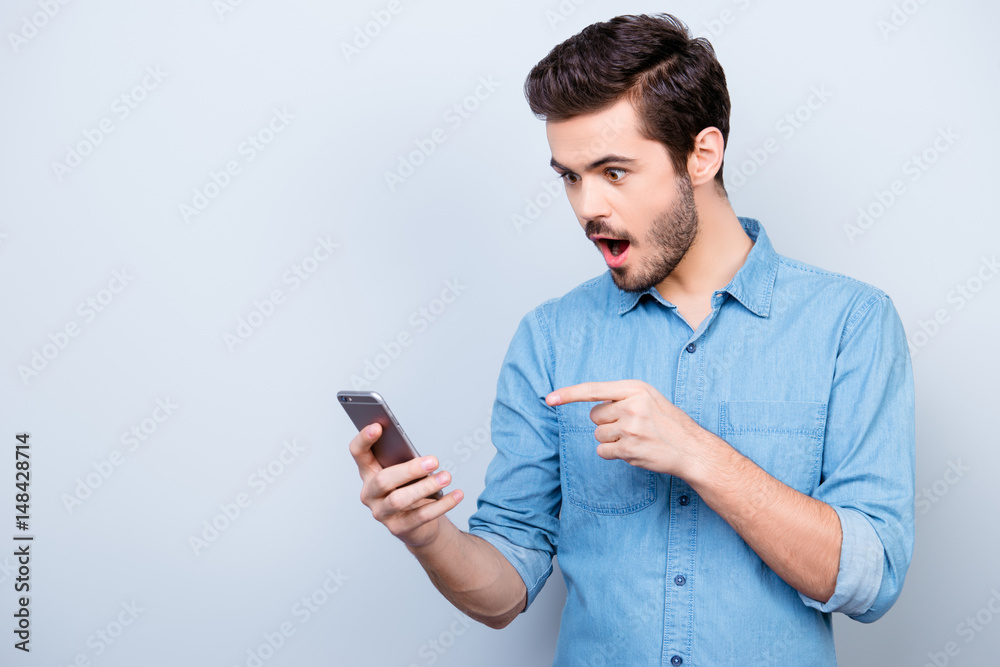Surprised young man is holding phone and reading message pointing on it with finger Surprised young man is holding phone and reading message pointing on it with finger