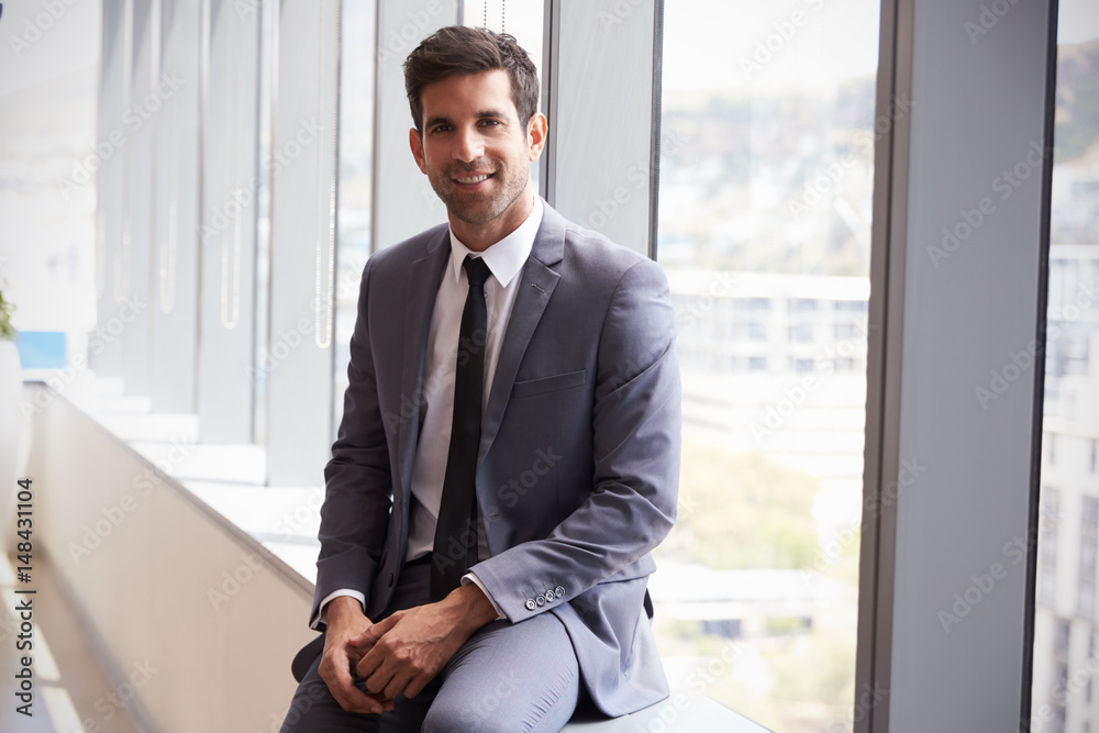 Portrait Of Young Businessman Sitting By Window In Office Portrait Of Young Businessman Sitting By Window In Office
