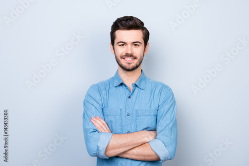 Close up portrait of cheerful brunet young man in jeans shirt crossing hands and look at camera on lifgt background
