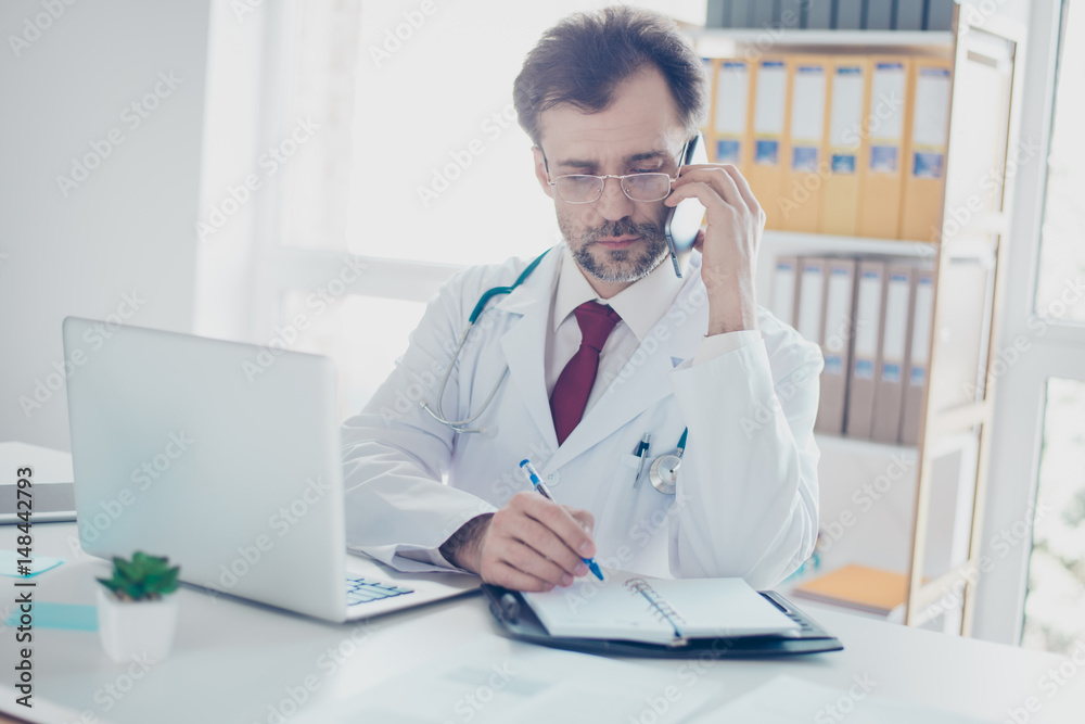 Serious doctor is consulting patient by the phone and writing down the details. He is in a white coat, glasses, sitting in modern office