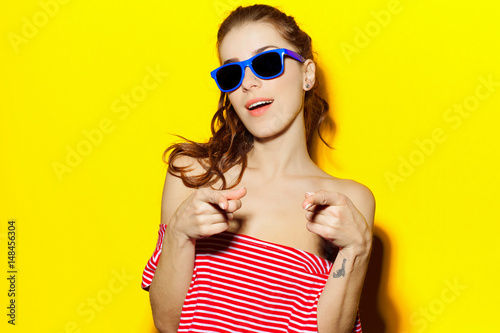 Beautiful young sexy girl in blue sunglasses and a red striped T-shirt laughing and having fun on a yellow background