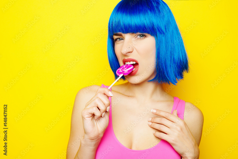 Beautiful young sexy girl in blue wig and pink bathing suit having fun and eating lollipop candy on yellow background Beautiful young sexy girl in blue wig and pink bathing suit having fun and eating lollipop candy on yellow background