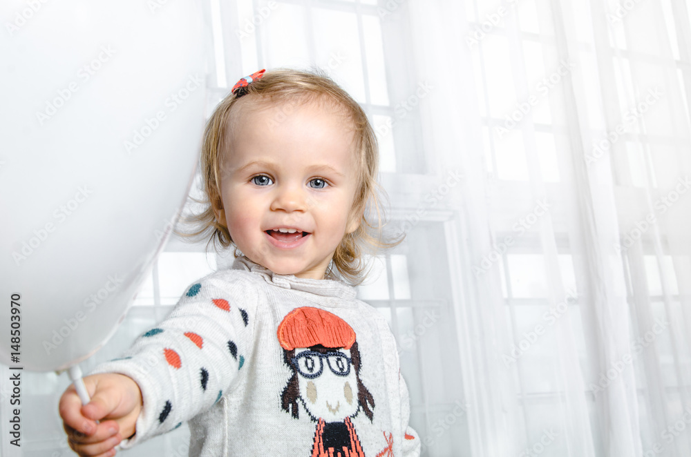 Close up little smiling girl baby on the background a child's room having fun and holding a white balloon Close up little smiling girl baby on the background a child's room having fun and holding a white balloon