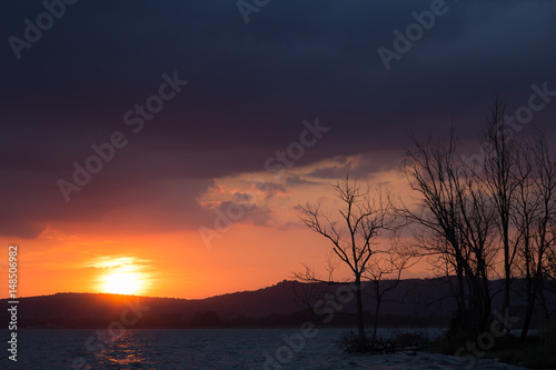 Sunset on a lake, with sun very low, dark and menacing clouds above, and some trees silhouettes in the foreground