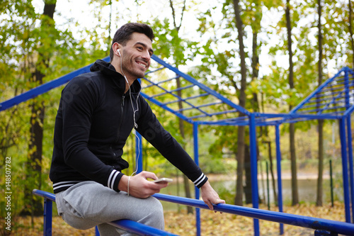 Cheerful young sporty man in headphones sitting on parallel bars in autumn park while taking short break during training